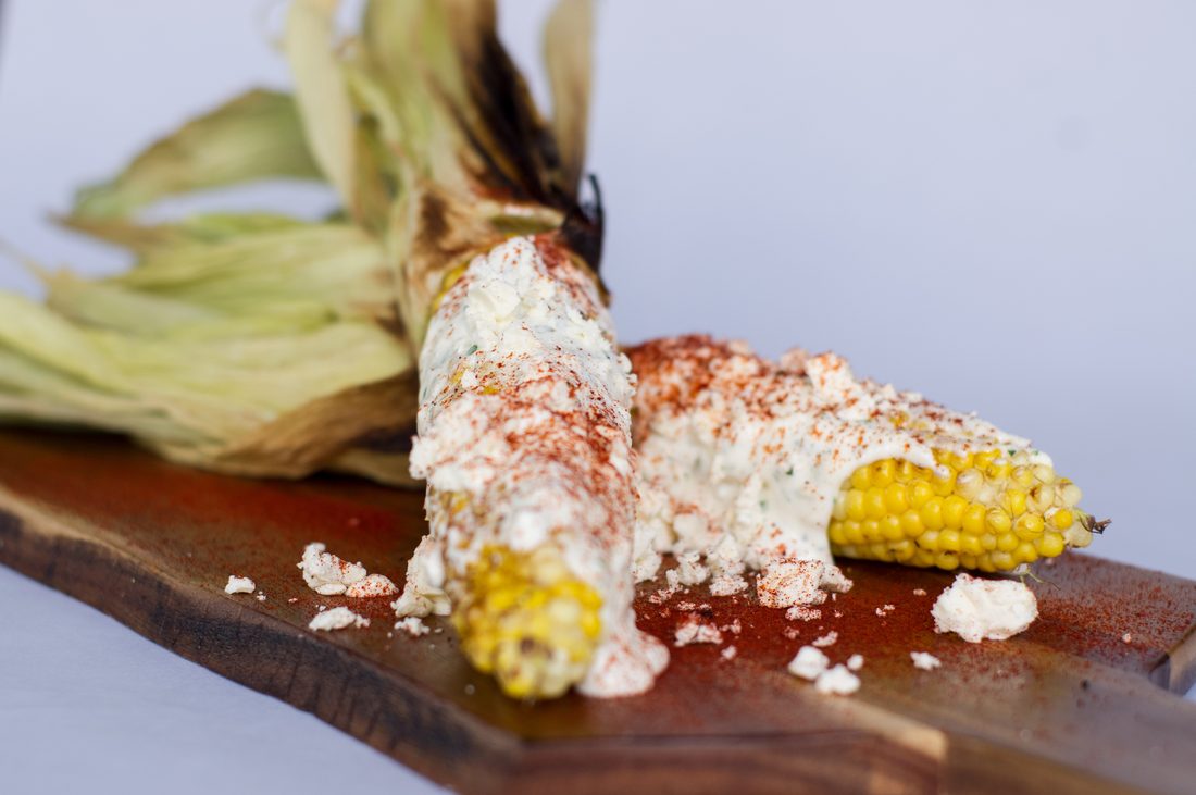 Grilled Mexican street corn on a wooden board, husk pulled back, coated in crema, crumbled queso fresco and chilli powder.