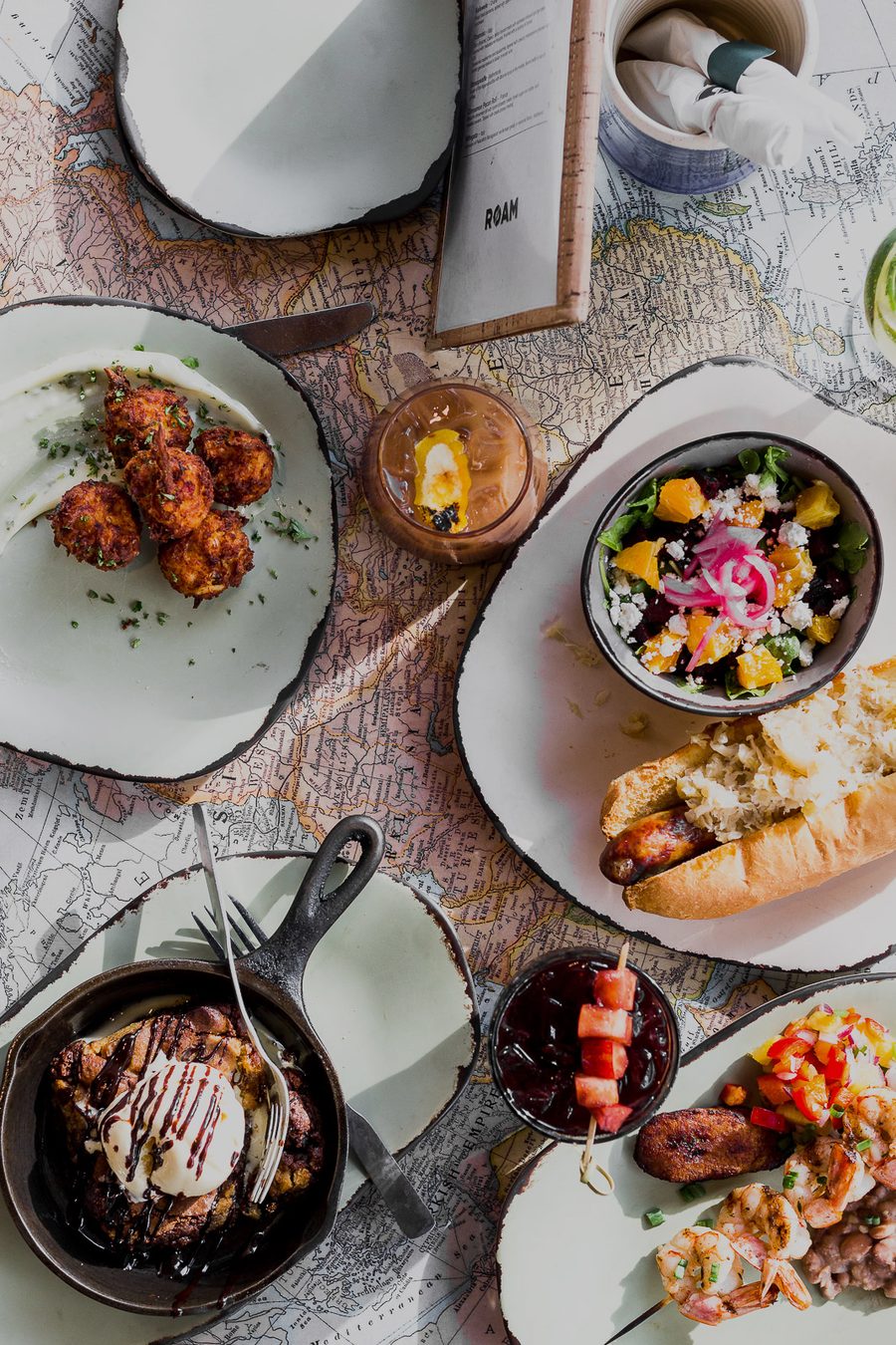 An overhead spread of ROAM dishes laid out on a vintage world map: fried hash balls, a citrus and beet salad, a sausage in a baguette, a cast iron skillet dessert, grilled shrimp and a ROAM menu card.