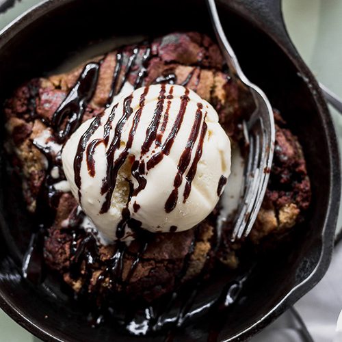 A warm skillet dessert topped with a scoop of vanilla ice cream and a chocolate drizzle, with a fork resting in the pan.