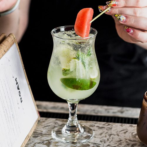 A tall ROAM cocktail with a mint and watermelon garnish, held at the bar by a server in a ROAM shirt.